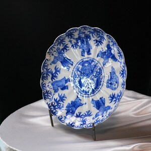 Puede incluir: Un plato de porcelana blanco y azul con borde festoneado. El plato presenta un dise&ntilde;o central y figuras en azul. El plato est&aacute; sostenido por un soporte met&aacute;lico, mostrando sus intrincados detalles y artesan&iacute;a.