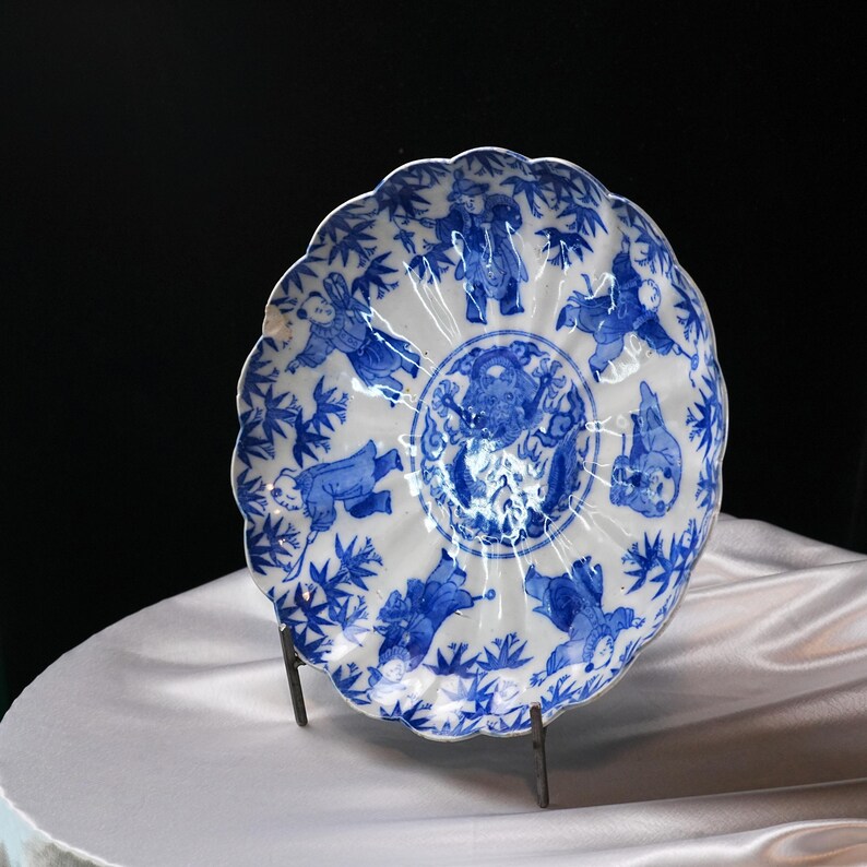 Puede incluir: Un plato de porcelana blanco y azul con borde festoneado. El plato presenta un dise&ntilde;o central y figuras en azul. El plato est&aacute; sostenido por un soporte met&aacute;lico, mostrando sus intrincados detalles y artesan&iacute;a.