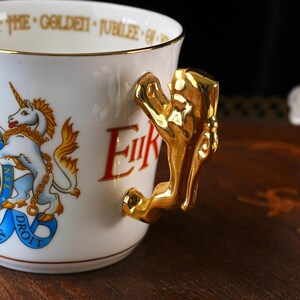 Puede incluir: Una taza de cer&aacute;mica blanca con un asa dorada en forma de le&oacute;n. El exterior muestra un emblema de unicornio colorido con una banda azul y el texto "DIEU ET MON DROIT". El borde interior tiene la inscripci&oacute;n "THE GOLDEN JUBILEE OF".