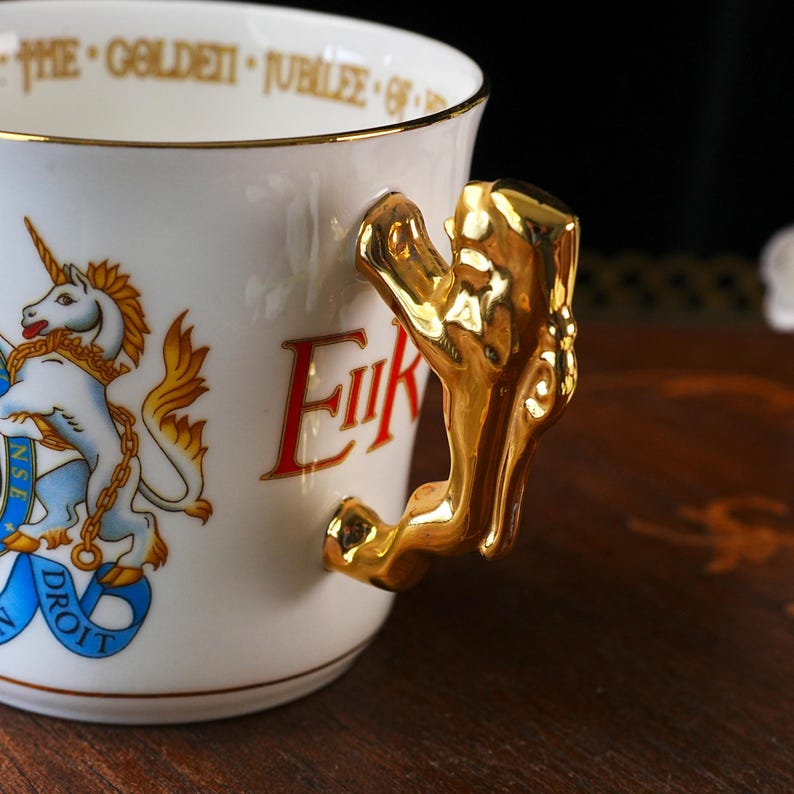 Puede incluir: Una taza de cer&aacute;mica blanca con un asa dorada en forma de le&oacute;n. El exterior muestra un emblema de unicornio colorido con una banda azul y el texto "DIEU ET MON DROIT". El borde interior tiene la inscripci&oacute;n "THE GOLDEN JUBILEE OF".