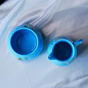 Op de afbeelding: Een felblauwe keramische suikerpot en melkkan set met een textuur op het oppervlak en gouden bladaccenten. Beide items hebben een gladde, diepblauwe binnenkant en worden van bovenaf getoond, waardoor de ronde openingen zichtbaar zijn.