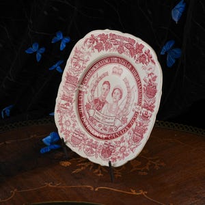 May include: A cream-coloured ceramic commemorative plate featuring a red transfer print of King George VI and Queen Elizabeth. The border displays provincial coats of arms and floral motifs with the text Commemorating the Visit of Their Majesties to the Dominion of Canada 1939.