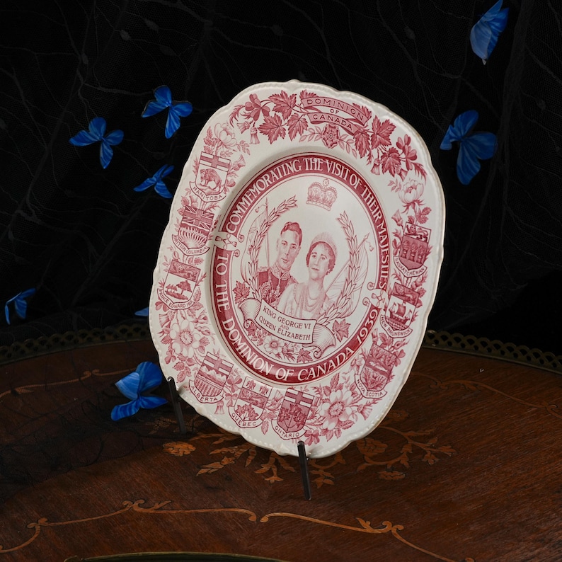 May include: A cream-coloured ceramic commemorative plate featuring a red transfer print of King George VI and Queen Elizabeth. The border displays provincial coats of arms and floral motifs with the text Commemorating the Visit of Their Majesties to the Dominion of Canada 1939.