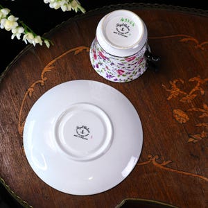 May include: A Royal Chelsea teacup and saucer set. The teacup, with a floral design in pink and purple, is upside down. The saucer is white. The set is on a dark wooden table with decorative inlay. Made in England.