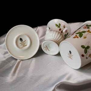 Op de afbeelding: Een collectie Wedgwood Wild Strawberry fijn porselein. De set bevat een schotel, een kleine voetbakschaal en een hoge cilindrische vaas. Elk stuk is wit met een aardbeien- en bladerpatroon.