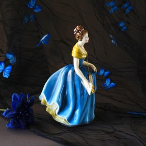 May include: A porcelain figurine of a person wearing a vibrant blue gown with a yellow shawl and a brown updo hairstyle. The figure holds a small yellow bouquet and wears a gold-toned pendant necklace. The dress features a full, flowing skirt.