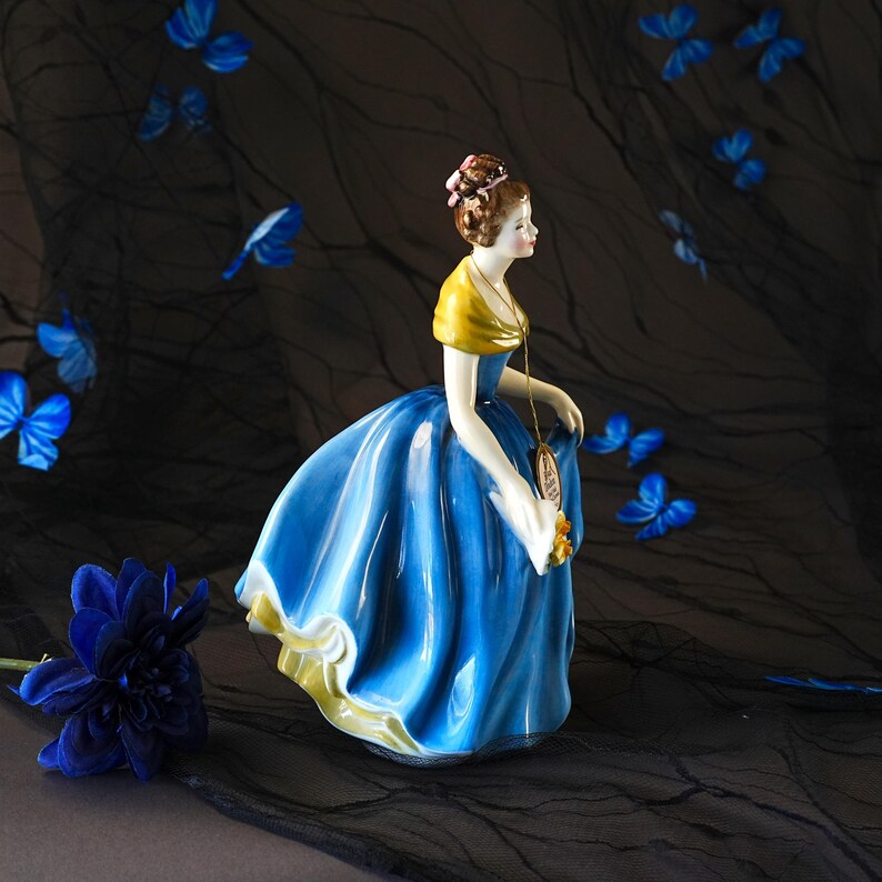 May include: A porcelain figurine of a person wearing a vibrant blue gown with a yellow shawl and a brown updo hairstyle. The figure holds a small yellow bouquet and wears a gold-toned pendant necklace. The dress features a full, flowing skirt.