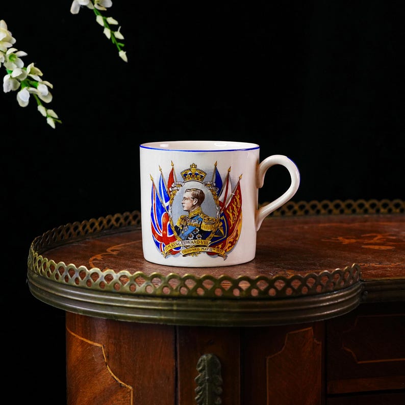 Op de afbeelding: Witte keramische mok met een blauwe rand, met een portret van koning Edward VIII, omringd door vlaggen en een kroon. De mok staat op een houten tafel met een decoratieve metalen rand.