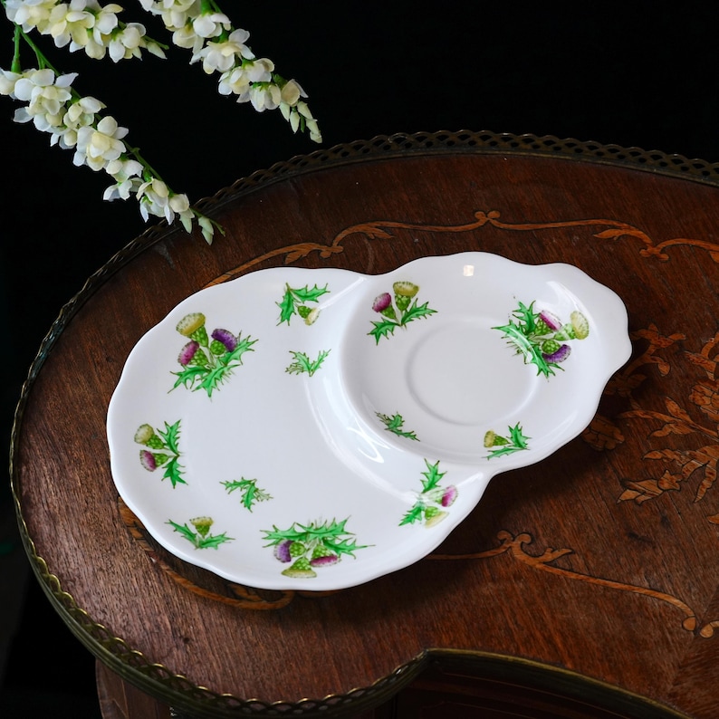 Puede incluir: Plato de cer&aacute;mica blanca con una forma &uacute;nica, con una hendidura circular central y una secci&oacute;n m&aacute;s grande y alargada. El plato est&aacute; decorado con dise&ntilde;os de cardos verdes y flores moradas. El plato est&aacute; sobre una superficie de madera.