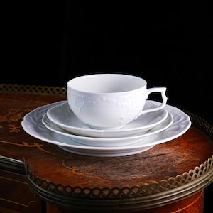 May include: A white porcelain teacup and saucer set with embossed floral detailing. The set includes a teacup, a small saucer, and a larger plate, all with scalloped edges. The teacup has a curved handle and sits atop the saucers on a wooden table.