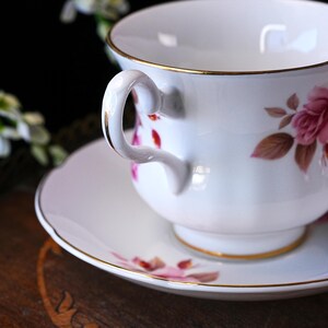 Op de afbeelding: Witte theekop en schotel set, versierd met roze en bruine bloemmotieven en gouden randen. De handgreep van de kop is wit met gouden details. De schotel heeft een geschulpte rand.