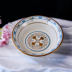 May include: A white ceramic bowl with a gold rim and a blue and gold decorative border. The bowl has a central design with floral and gold accents, and vertical ribbed detailing.