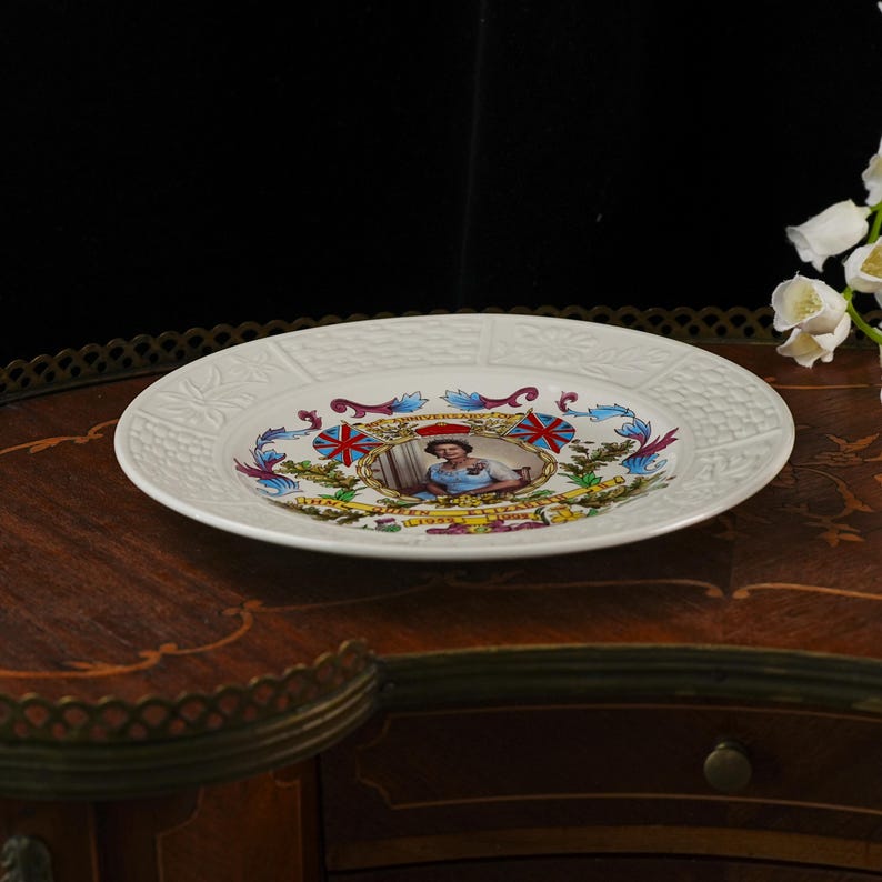 Op de afbeelding: Een wit keramieken herdenkingsbord met een centraal portret van koningin Elizabeth II, geflankeerd door twee Union Jack-vlaggen en bloemmotieven. De tekst luidt 40th Anniversary en Queen Elizabeth 1952-1992. De rand heeft een reli&euml;fpatroon.