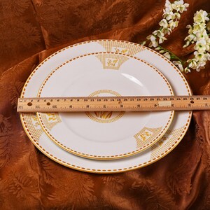 Op de afbeelding: Twee witte keramische borden met een gouden bloemenontwerp en gouden rand. Een houten liniaal ligt over de borden en meet ongeveer 30 cm. De borden liggen op een bruin oppervlak met een patroon.