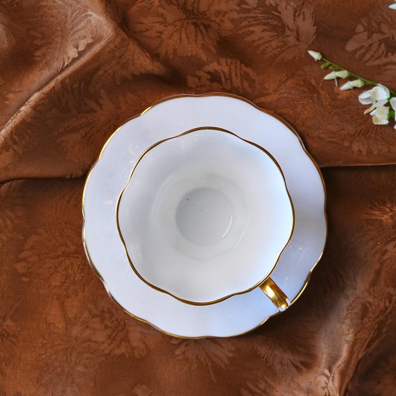 May include: A white teacup and saucer set with gold trim. The cup and saucer have a scalloped edge. The set is placed on a brown patterned fabric. A sprig of white flowers is in the background.