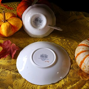 May include: A white teacup and saucer set with gold trim, featuring the "Paragon" brand. The set is placed on a yellow lace tablecloth, with pumpkins and a red leaf in the background. The teacup is upside down.