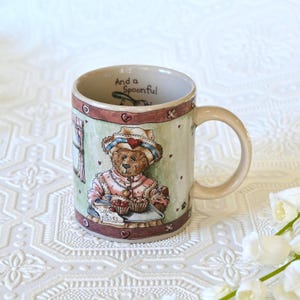 Puede incluir: Taza de cerámica beige con un diseño de oso de peluche. El oso lleva un sombrero y sostiene una bandeja de cupcakes. La taza tiene las palabras "And a Spoonful" y "Love Kisses". La taza tiene un asa curva.