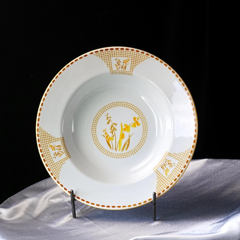 May include: White ceramic soup bowl with a yellow floral design. The bowl features a yellow and brown checkered border and a central floral motif. The rim is outlined with a dotted brown line. The bowl is displayed on a black metal stand.