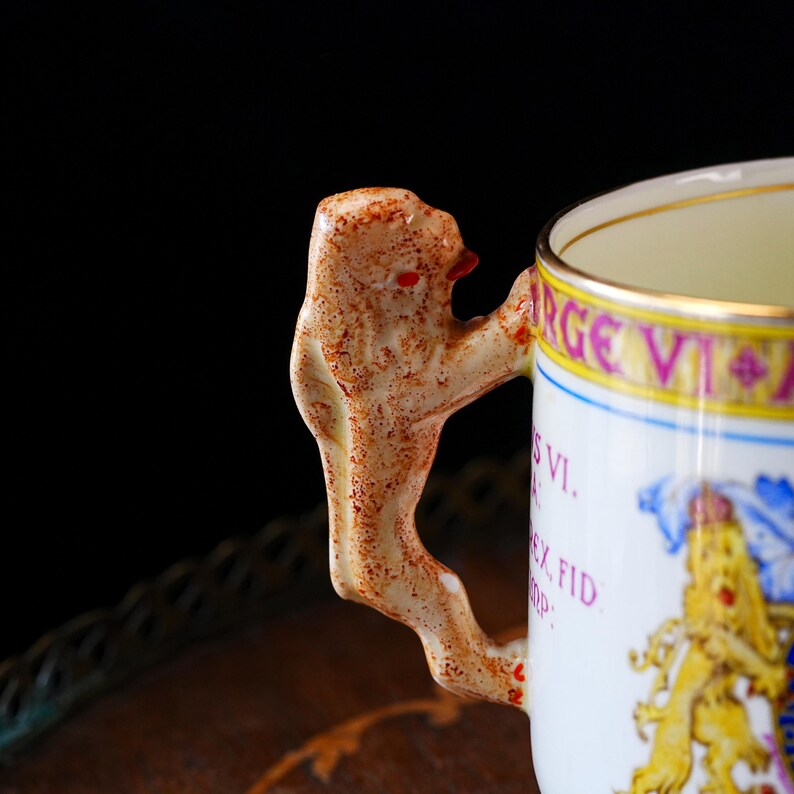 Puede incluir: Taza de cer&aacute;mica blanca con un asa decorativa en forma de le&oacute;n. La taza presenta un borde dorado y texto en rosa y azul. El asa del le&oacute;n es beige con detalles rojos. La taza es probablemente vintage o antigua.