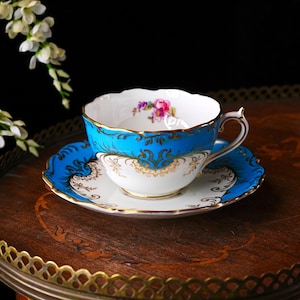 Op de afbeelding: Een vintage kop en schotel set met een levendig turquoise en goud design. De theekop heeft een bloemenpatroon aan de binnenkant en een delicate oor. De set staat op een decoratieve houten tafel.