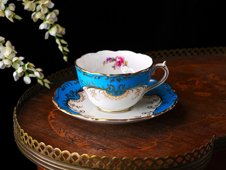 Op de afbeelding: Een vintage kop en schotel set met een levendig turquoise en goud design. De theekop heeft een bloemenpatroon aan de binnenkant en een delicate oor. De set staat op een decoratieve houten tafel.