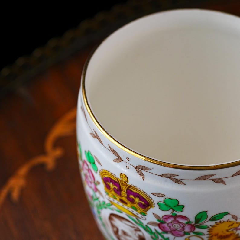 Może przedstawiać: Biały porcelanowy kubek ze złotym obrzeżem i ozdobnym paskiem z brązowych liści. Kubek posiada kolorowy emblemat z koroną, r&oacute;żowymi kwiatami, zielonymi koniczynami i lwem, umieszczony na gładkiej, błyszczącej białej powierzchni.