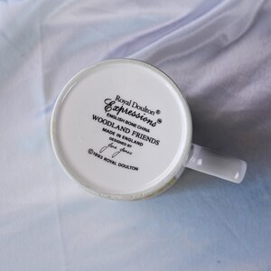 Puede incluir: La parte inferior de una taza de porcelana de hueso blanca con texto impreso en negro que dice Royal Doulton Expressions English Bone China WOODLAND FRIENDS MADE IN ENGLAND DESIGNED BY Jane Jones 1993 ROYAL DOULTON con un asa a la derecha.