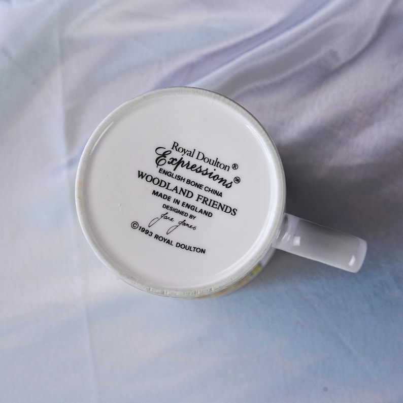 Puede incluir: La parte inferior de una taza de porcelana de hueso blanca con texto impreso en negro que dice Royal Doulton Expressions English Bone China WOODLAND FRIENDS MADE IN ENGLAND DESIGNED BY Jane Jones 1993 ROYAL DOULTON con un asa a la derecha.
