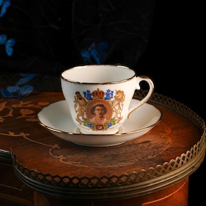May include: A white porcelain teacup and saucer set featuring a gold-trimmed rim and handle. The cup displays a central portrait of Queen Elizabeth II, flanked by a lion and unicorn crest with a crown above and the text Coronation June 2nd 1953.