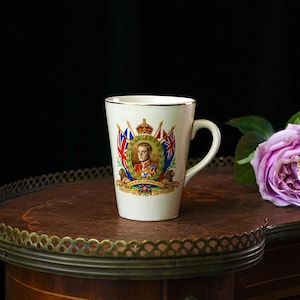 Puede incluir: Taza de cer&aacute;mica color crema con borde dorado, con un retrato del rey Eduardo VII, banderas y el texto "Coronation May 12th 1937". La taza est&aacute; sobre una mesa de madera con borde decorativo, con una rosa p&uacute;rpura en el fondo.