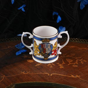 May include: A white porcelain two-handled mug featuring a central royal coat of arms with a crown, a shield, and two lions. The design includes a blue border with gold trim and the text HONI SOIT QUI MAL Y PENSE inscribed around the shield.