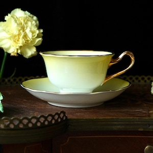 Könnte beinhalten: Ein Vintage-Tee-Tassen- und Untertassen-Set. Die Tasse und Untertasse sind hellgelb mit goldenem Rand. Der Griff der Tasse ist goldfarben. Das Set steht auf einer dunklen Holzoberfläche, mit einer gelben Blume im Hintergrund.