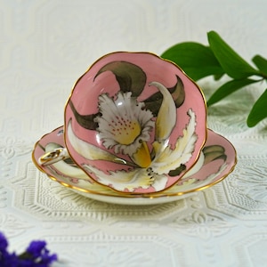 May include: A pink teacup and saucer set with a floral design. The teacup and saucer are painted with a white and yellow flower with green and brown accents. The rims are trimmed with gold.