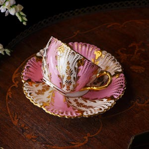 May include: A pink and white teacup and saucer set with gold accents. The teacup has a gold handle and is tilted on the matching saucer. The set features a scalloped edge and ornate gold floral designs. A classic tea set.