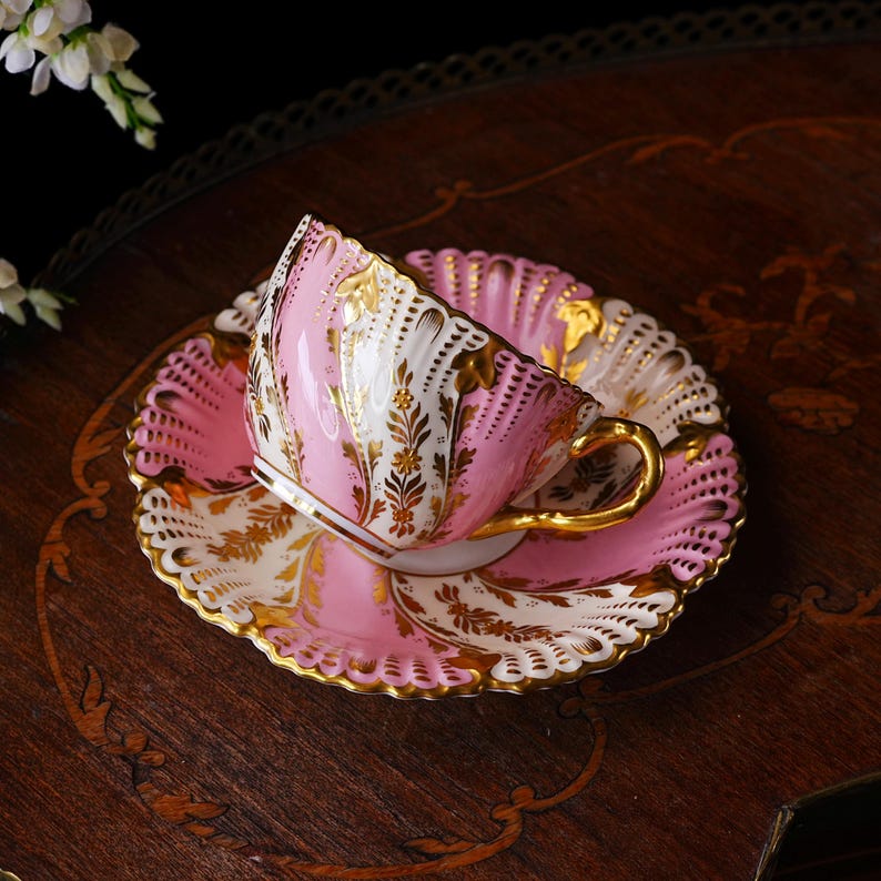 May include: A pink and white teacup and saucer set with gold accents. The teacup has a gold handle and is tilted on the matching saucer. The set features a scalloped edge and ornate gold floral designs. A classic tea set.