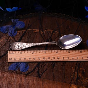 Puede incluir: Una cuchara de metal plateado con un dise&ntilde;o en relieve de flores y un ave en el mango, colocada sobre una regla de madera que muestra una longitud de aproximadamente 14,6 cm desde el extremo del mango hasta el borde de la cuchara.