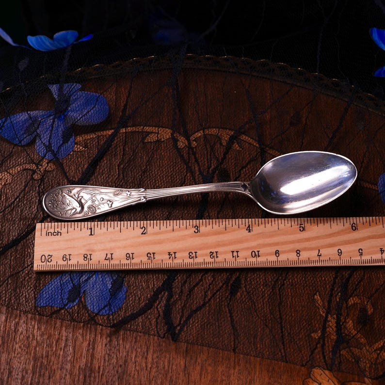 Puede incluir: Una cuchara de metal plateado con un dise&ntilde;o en relieve de flores y un ave en el mango, colocada sobre una regla de madera que muestra una longitud de aproximadamente 14,6 cm desde el extremo del mango hasta el borde de la cuchara.