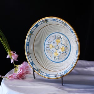 May include: A white ceramic plate with a gold rim and blue and gold decorative accents. The plate features a central design with gold and blue floral motifs. The plate is displayed on a black metal stand, with pink flowers in the background.