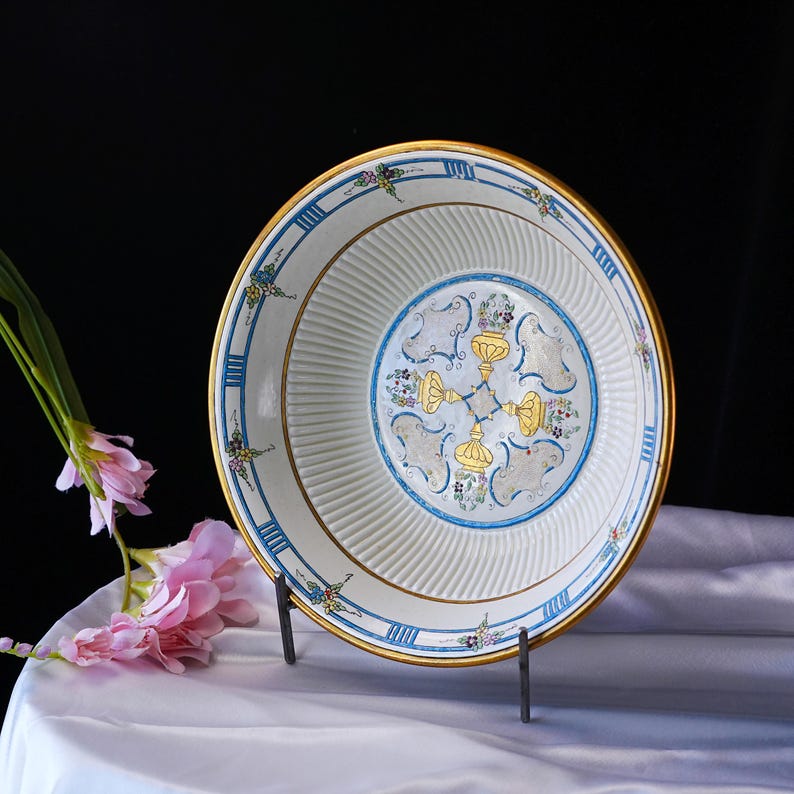 May include: A white ceramic plate with a gold rim and blue and gold decorative accents. The plate features a central design with gold and blue floral motifs. The plate is displayed on a black metal stand, with pink flowers in the background.