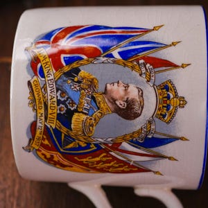 Op de afbeelding: Witte keramische mok met een portret van koning Edward VIII, gekroond op 12 mei 1937, met de Union Jack. De mok heeft een blauwe rand en oor, met gouden accenten en tekst.