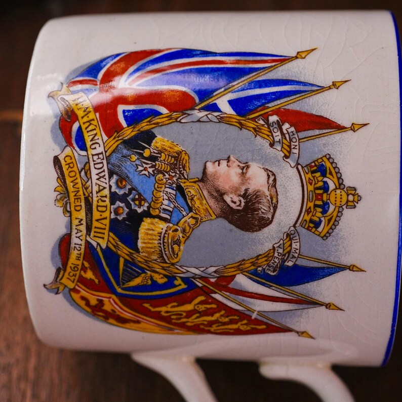 Op de afbeelding: Witte keramische mok met een portret van koning Edward VIII, gekroond op 12 mei 1937, met de Union Jack. De mok heeft een blauwe rand en oor, met gouden accenten en tekst.