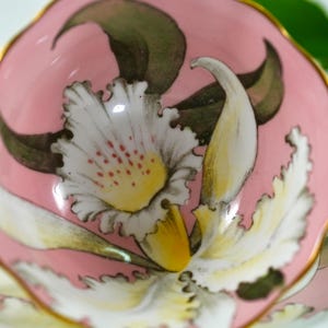 May include: A small, pink ceramic dish with a floral design. The design features a large white and yellow flower with red accents, surrounded by green leaves. The rim is trimmed with gold.