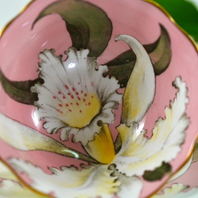 May include: A small, pink ceramic dish with a floral design. The design features a large white and yellow flower with red accents, surrounded by green leaves. The rim is trimmed with gold.