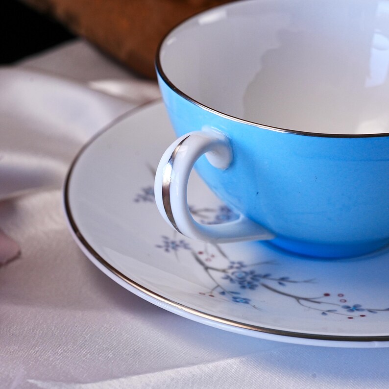 Pode incluir: Uma ch&aacute;vena de ch&aacute; azul claro com interior branco e pires a condizer. A ch&aacute;vena e o pires t&ecirc;m uma borda prateada. O pires apresenta um delicado padr&atilde;o floral em azul e vermelho. A pega da ch&aacute;vena &eacute; branca com um detalhe prateado.