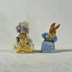 以下が含まれることがあります： 2つの陶器製の擬人化された動物の置物。1つはクリーム色のドレスと帽子を着て鏡を持っているネズミです。もう1つは青いドレスを着て黄色い物を持っている茶色のウサギです。どちらも塗装されたディテールがあります。