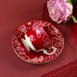 May include: A red teacup and saucer set with white leaf patterns and gold trim. The teacup is resting on the saucer, with a pink rose in the background. The set is designed for tea parties and special occasions.