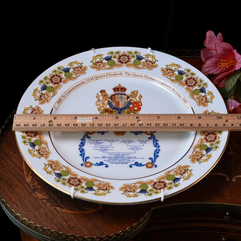 Pode incluir: Um prato comemorativo de porcelana branca com um bras&atilde;o real e uma &aacute;rvore geneal&oacute;gica, com uma r&eacute;gua de madeira no centro mostrando um di&acirc;metro de 25 cm. A borda &eacute; decorada com motivos florais coloridos, incluindo cardos e rosas.