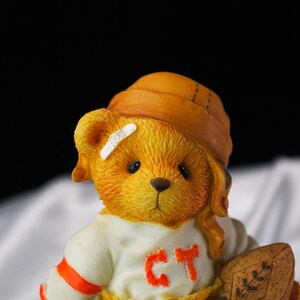 Op de afbeelding: Gedetailleerd beeldje van een teddybeer met een bruine pet en een wit shirt met de letters "CT" in oranje. De beer heeft een pleister op zijn hoofd en houdt een voetbal vast. De beer is lichtbruin.
