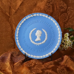 May include: A blue commemorative plate with white relief detailing. The plate features a profile of Queen Elizabeth II within a laurel wreath, with the text "Silver Jubilee 1952-1977". A decorative item for collectors.