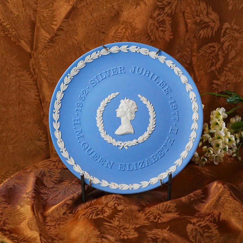 May include: A blue commemorative plate with white relief detailing. The plate features a profile of Queen Elizabeth II within a laurel wreath, with the text "Silver Jubilee 1952-1977". A decorative item for collectors.